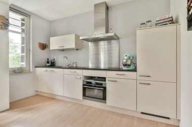 Gösterişli, modern bir mutfak, minimalist dolaplar, paslanmaz çelik aletler, ve geniş pencereden yayılan geniş doğal ışık..