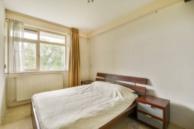 Geniş ve modern bir yatak odası, hafif yorganı olan bir yatak odası, doğal ışığa izin veren bir pencere ve ahşap bir komodin. Duvarlar tarafsız tonlarda boyanmış..