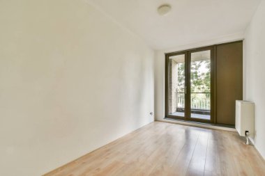 Parlak, minimalist bir odada ahşap zemin ve geniş pencereler var. Balkon manzarasına açık, açıklık ve sükunet hissi veriyor..