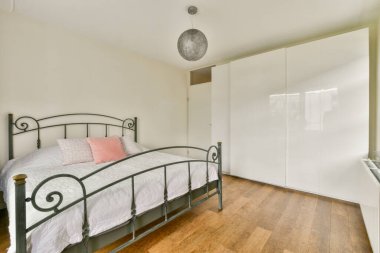 Parlak ve havadar minimalist bir yatak odası. İçinde metal bir yatak çerçevesi, beyaz bir gardırop, ahşap döşeme, iç tasarımda sadelik ve zarafet var..