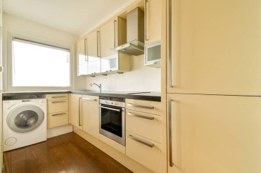 Parlak, modern bir mutfak. İçinde bej dolaplar, paslanmaz çelik aletler ve camdan içeri ışık saçan geniş bir düzen var..