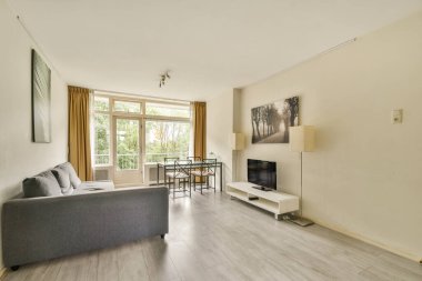 Gri bir kanepe, minimalist bir televizyon ünitesi ve doğal ışığın boşluğu doldurmasına izin veren geniş pencereli şık bir oturma odası..