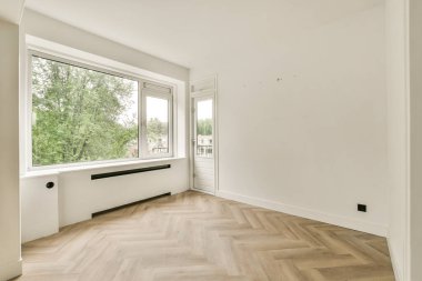 Geniş, minimalist bir iç mekan. Doğal ışık sağlayan geniş bir pencere. Odada açık ahşap bir zemin ve çıplak beyaz duvarlar var..