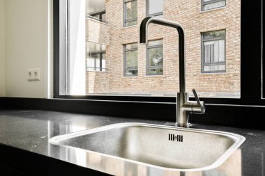 Modern tuğla bir binaya bakan büyük bir pencerenin yanında şık bir mutfak lavabosu. Ayar, şehir estetiğiyle çağdaş tasarımı birleştiriyor..