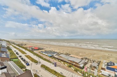 Panoramik bir manzara kumlu bir plajı, okyanus dalgalarını ve parlak bir gökyüzünün altında renkli deniz kenarındaki yapılarla dolu canlı bir tahta kaldırımı gösteriyor..