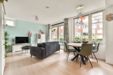 Rahat bir kanepe, yemek masası ve doğal ışık sağlayan geniş pencereli modern bir oturma odası, çağdaş mobilyalar ve dekorlar sergileniyor..