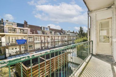 Mavi gökyüzünün altında bulutlu bir sıra şehir binasını gören korkulukların olduğu bir balkon manzarası. Yeşillik doğanın bir dokunuşunu ekler..