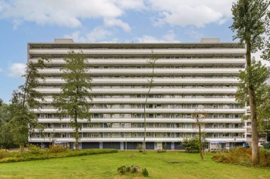 Parlak çizgileri ve açık balkonlarıyla karakterize edilen çağdaş çok katlı bir bina, ağaçlarla dolu yemyeşil bir araziye karşı kurulmuş..