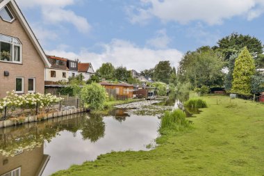 Bereketli yeşillik ve büyüleyici evlerle dolu sakin bir rıhtım manzarası sergileniyor, dingin bulutları ve canlı doğayı yansıtıyor..