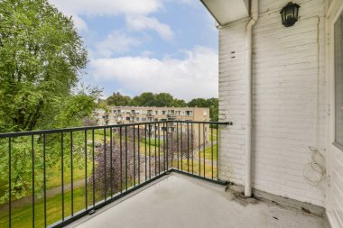 Huzurlu bir balkon manzarası, bulutlu gökyüzünün altında modern bir yerleşim zeminiyle tamamlanmış gür ağaçlar ve canlı yeşillikler içerir. Doğa aşıkları için mükemmel..