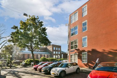 Canlı bir şehir manzarası. Tuğla bina, park edilmiş arabalar ve açık gökyüzünün altında yemyeşil ağaçlar. Şehir yaşamını resmediyor..