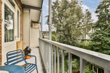 Huzurlu bir balkon sahnesinde mavi sandalyeler ve yemyeşil ağaçların huzurlu manzarası ve arka planda rahatlatıcı bir yapı yer alıyor..