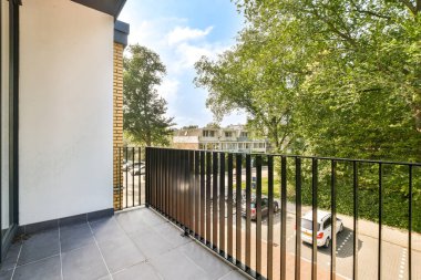 Ağaçlara ve park edilmiş arabalara bakan çağdaş bir balkon kentsel ve doğal elementlerin bir karışımını sergiliyor. Mekan, parlak güneş ışığıyla davetkar ve huzurlu..