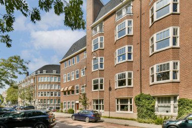 Geniş pencereli ve eşsiz tasarım özelliklerine sahip zarif modern binaların sergilendiği bir manzara, yeşillikle dolu canlı bir şehir ortamında geçiyor..