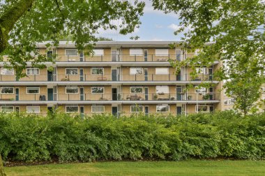 Çağdaş bir konut binasında birden fazla balkon ve geniş pencereler, ön planda yemyeşil ile harmanlanmış, kentsel yaşamı gösteren.