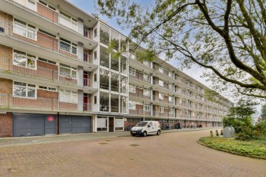 Sakin yeşillik ve sokak unsurları ile çevrili geniş pencere ve balkonların yer aldığı modern bir bina, kentsel yaşamı gözler önüne seriyor..