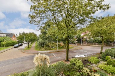 Parçalı bulutlu bir gökyüzünün altında bakımlı ağaçlar, bahçeler ve caddelerin yer aldığı sakin bir mahalle manzarası huzurlu bir atmosfer yaratıyor..