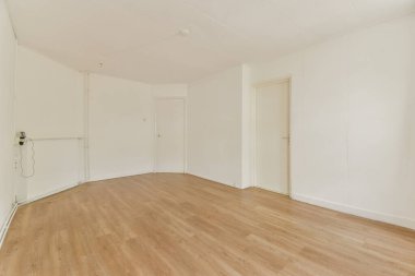 Boş, aydınlık bir iç oda. Açık ahşap döşeme ve sade beyaz duvarlar sergileniyor. Temiz ve minimalist bir estetik sunuyor..