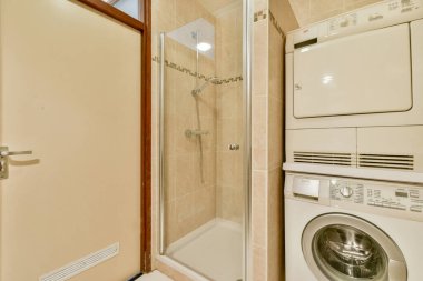 Cam duş bölmesi olan küçük ve çağdaş bir banyo. Çamaşır birimlerini istifleyip temiz ve verimli bir düzen sergiliyor..
