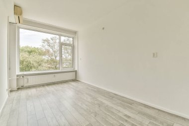 Geniş, dağınık bir oda. Geniş pencereler var. Alanı doğal ışıkla dolduran, soluk ahşap zemin ve tarafsız duvarlarla donatılmış..