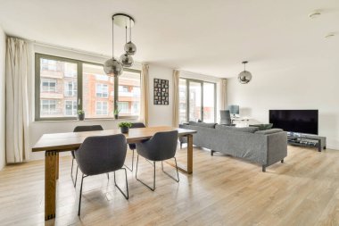 Şık ve çağdaş bir oturma odası, ahşap bir yemek masası, rahat koltuklar, ve geniş pencereler, boşluğa doğal ışık girmesini sağlıyor..