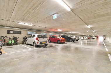 Geniş ve iyi ışıklandırılmış bir yeraltı otoparkı. Belirlenmiş noktalarda düzenli olarak park edilmiş araçlar ve bisikletlerin yer aldığı..