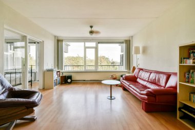 Parlak ve ferah bir modern oturma odası kırmızı deri bir kanepe, bir sehpa, ve doğal ışık sağlayan büyük pencereler ve dışarıda bir manzara..