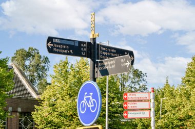 Çoklu yön işaretleri, açık bir gökyüzü altında, çevre bölgelere döngü sembolleri ve mesafeler içeren çeşitli rotaları ve konumları gösterir..
