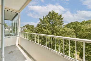 Geniş, modern bir balkon. Pürüzsüz bir korkuluk ve yemyeşil ağaçlar ve mavi gökyüzü. Huzurlu bir açık hava alanı yaratıyor..