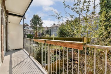 Tahta korkuluklar ve bol yeşillik içeren sakin bir balkon sahnesi, rahatlama ve doğa takdiri için ideal. Güneş ışığı yere gölge düşürür..