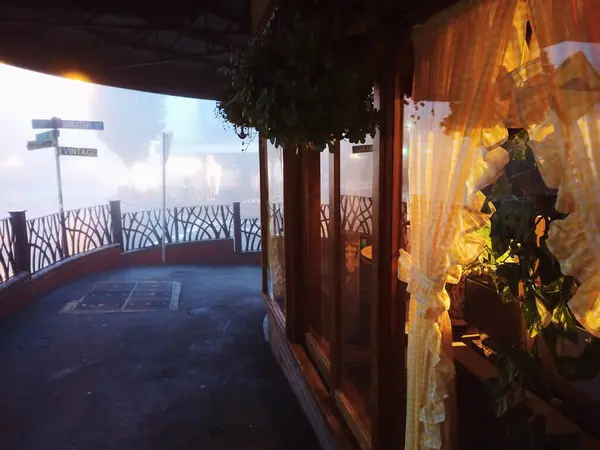 Stock image A cozy view from a warmly lit window with curtains, overlooking a foggy street corner. The mist envelops the town, creating a serene and nostalgic atmosphere