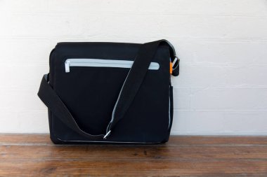 Ön fermuarı ve ayarlanabilir omuz kayışı olan parlak siyah bir kurye çantası, tahta bir yüzeyde beyaz tuğla bir duvara karşı sergileniyor. Çantanın içinde minimalist, modern bir tasarım var.