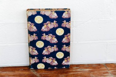 A vintage book with a blue fabric cover featuring a repetitive pattern of intricate, bag-like designs and golden round emblems. The book is set against a white wall and rests on a wooden surface, showcasing its detailed design clipart