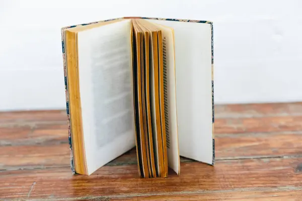 stock image An open vintage book displayed upright, revealing yellowed pages and a patterned hardcover. The book is set on a wooden surface against a white wall, highlighting its age and texture