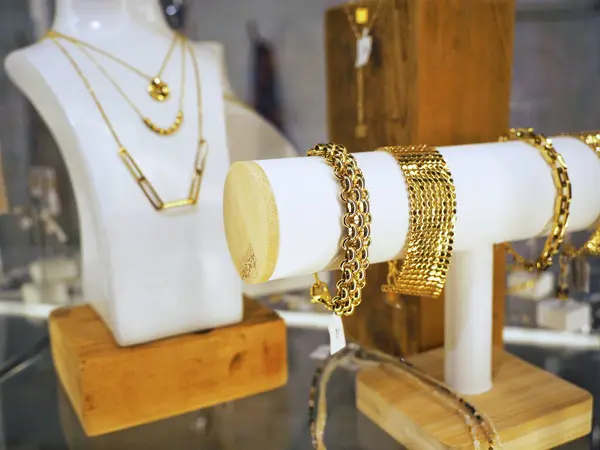 Ahşap tabanlı beyaz stantlara yerleştirilmiş zarif altın kolyeler ve bileziklerin yer aldığı bir butik mücevher sergisinin yakın çekimi. Parçalar lüks ve tarz yayıyor.