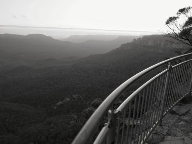 Kıvrımlı bir metal korkuluk geniş bir orman vadisini, katmanlı tepeleri, uçurumları ve Avustralya 'nın vahşi doğasının bu sakin siyah beyaz fotoğrafında puslu bir gökyüzünü çerçeveler..