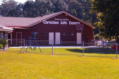 A fenced playground with a swing set in front of the Nambucca Valley Christian Life Centre building, surrounded by greenery and trees on a sunny day. clipart