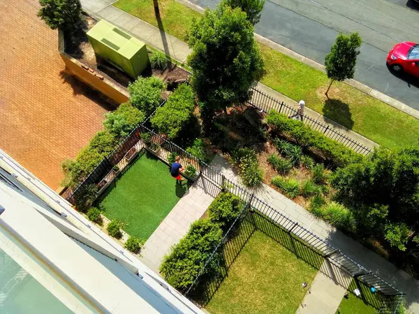 Yeşil çimleri, patikaları ve çalıları olan bir konut bahçesinin yüksek açılı görüntüsü, yan kaldırımda bir kişi ve bir yayayı gösteriyor..