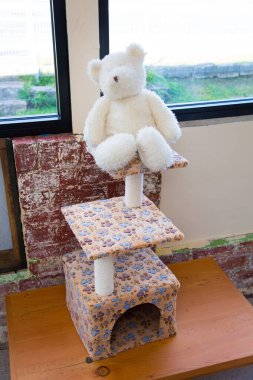 A white teddy bear placed on a multi-level cat scratching post with paw-print fabric, set against a rustic brick wall and a window overlooking greenery. clipart