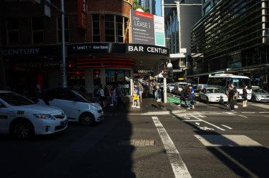 George Street 'in Sydney' deki editör görüntüsü, yayaların kalabalık bir kavşaktan geçişlerini, taksilerin ve otobüslerin trafiğini, ve öğleden sonra güneşinin altındaki hareketli mağazaları yakalıyor.