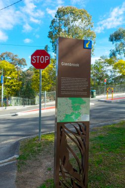 Glenbrook, Blue Mountains 'a gelen ziyaretçileri karşılayan, içinde bir harita, karmaşık bir tasarım ve yakındaki dur levhası bulunan, ağaçların ve güneşli bir banliyö sokağının olduğu bir bilgilendirme tabelası.