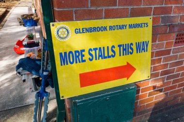 A vibrant yellow directional sign for the Glenbrook Rotary Markets, pointing visitors toward additional market stalls, mounted on a brick wall in a sunny outdoor setting clipart