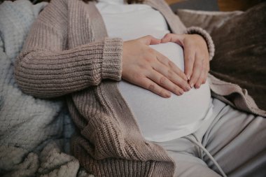 Mother to be heavily pregnant sitting on the sofa making heart shape symbol on belly. High quality photo