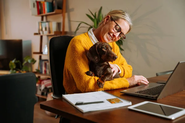 在笔记本电脑上边打字边拿着宠物狗在家工作的高加索自由职业者 高质量的照片 — 图库照片
