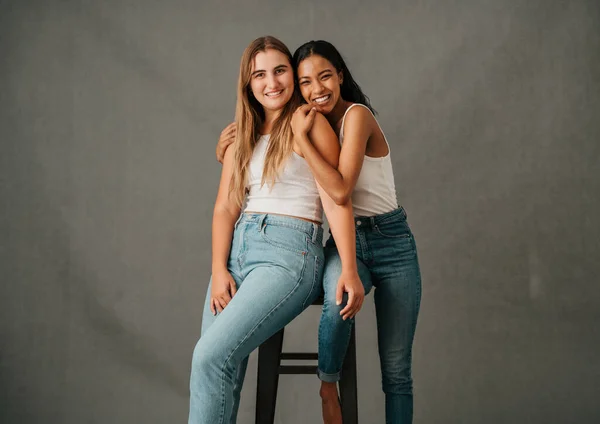 Two girls friends smiling happily sharing high a stool in there jeans and t shirts. High quality photo