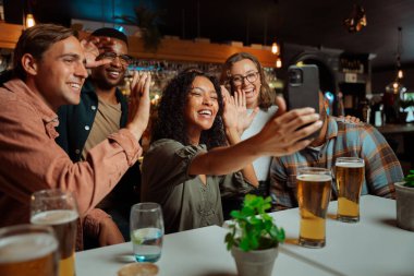 Çok kültürlü bir arkadaş grubu restoranda oturmuş arkadaşlarıyla video görüşmesi yaparken kameraya el sallıyorlar. Yüksek kalite fotoğraf