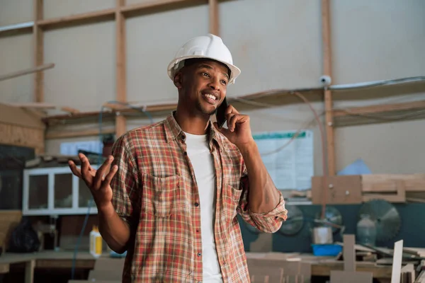 African American male wears hardhat while discussing business on phone in carpentry warehouse . High quality photo
