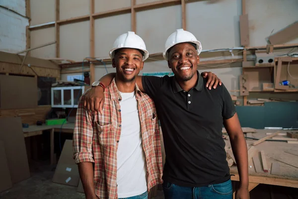 African American colleagues wearing hardhats stand with arms around each other in warehouse . High quality photo
