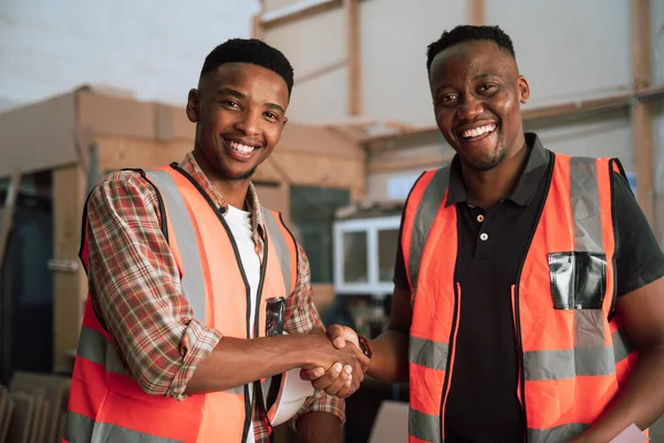 Smiling African American colleagues wearing safety vests shake hands in woodwork factory . High quality photo