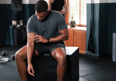 Şapka takan bir adam spor salonundaki bir bankta oturmuş saatlerini izliyor. Bileğini kontrol ederken bacak kasları görünür durumda.